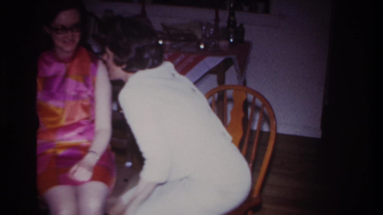 A woman in a light-colored dress leans in toward a seated woman wearing a vivid 1960s patterned dress inside a private home.