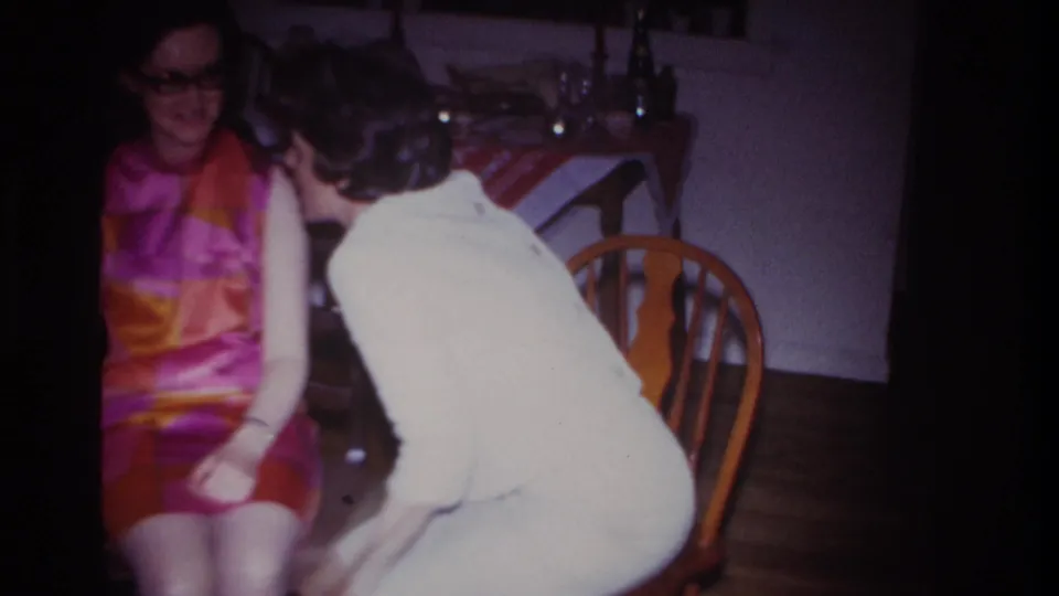 A woman in a light-colored dress leans in toward a seated woman wearing a vivid 1960s patterned dress inside a private home.