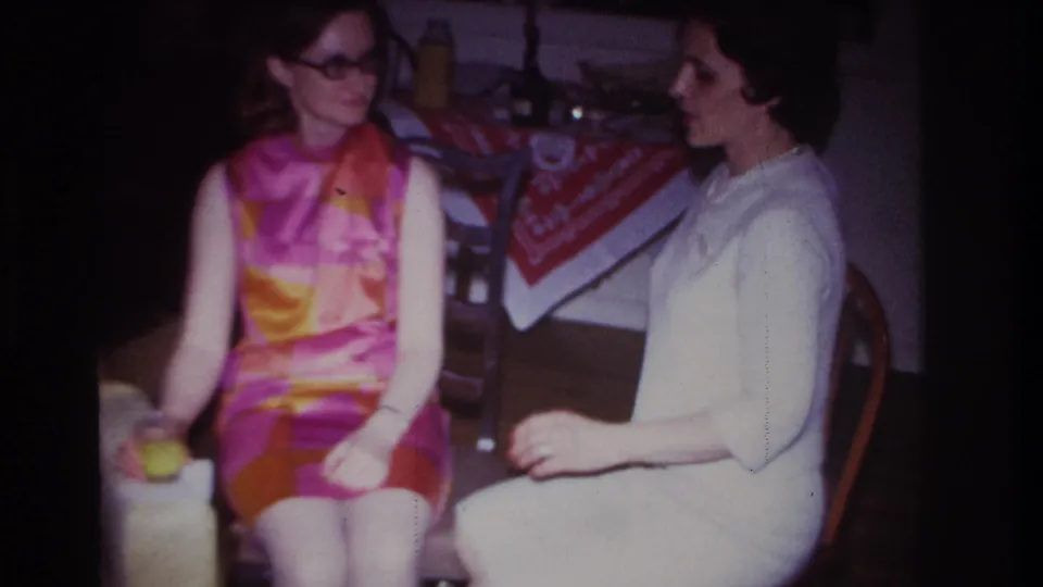 Two adult women face each other in a 1967 Rockaway Beach home interior, one holding a small green object near her lap.