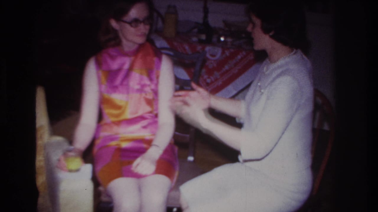 Two seated women converse indoors, one in a bright sleeveless dress and one in a white dress.