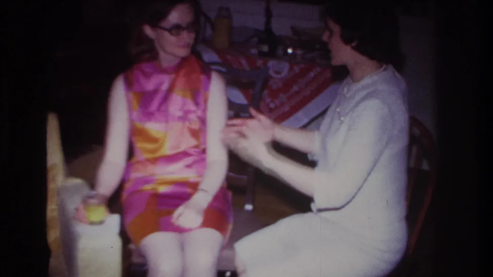 Rockaway Beach 1967 home-movie frame of two women seated indoors, with a bright geometric dress, white dress, drink glass, and tablecloth behind them.
