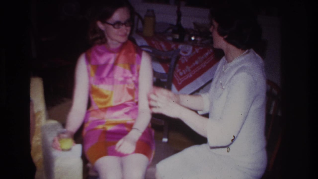 1967 8mm frame from Rockaway Beach showing a woman in white gesturing toward a seated woman in a vivid pink, orange, and yellow dress.