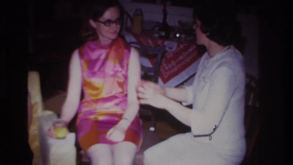 1967 8mm frame from Rockaway Beach showing a woman in white gesturing toward a seated woman in a vivid pink, orange, and yellow dress.