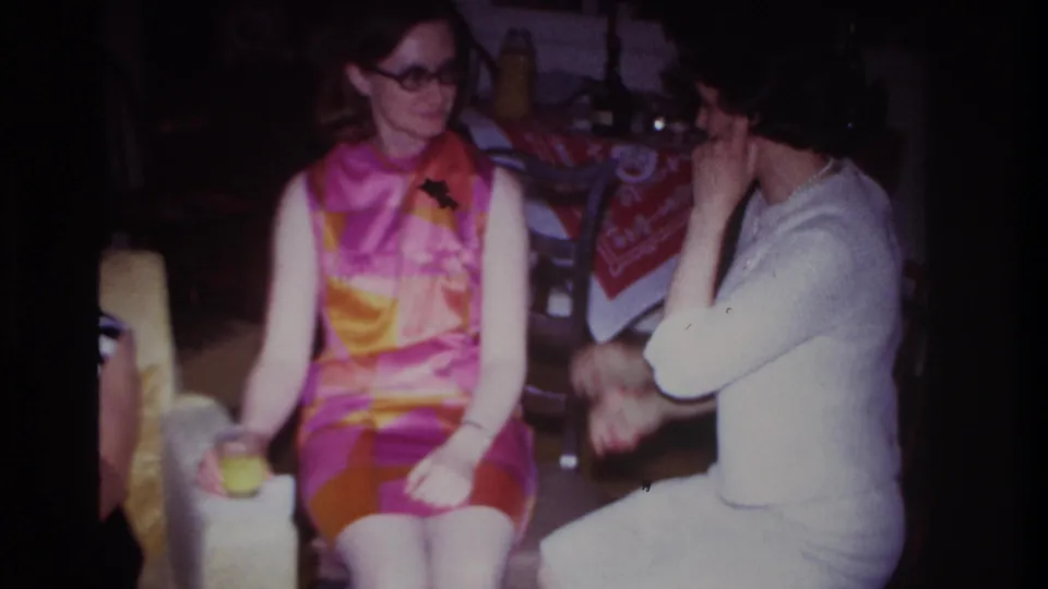 Rockaway Beach 1967 home-movie frame with two seated women indoors, the woman in white turned partly away and the bright dress centered.
