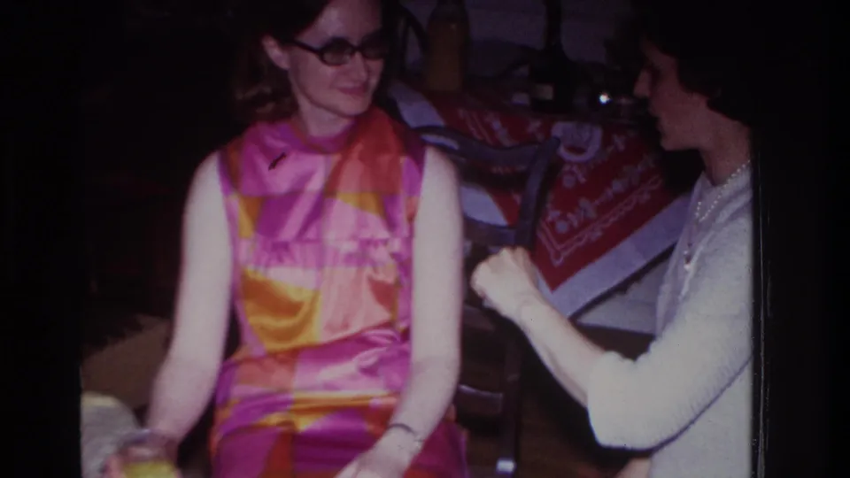 1967 Rockaway Beach 8mm frame of two women seated indoors, one smiling in a colorful dress while the other sits near a red patterned tablecloth.