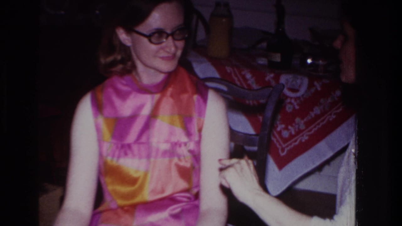 In 1967 Rockaway Beach home-movie footage, a young woman in glasses and a bright sleeveless dress sits at a patterned table.
