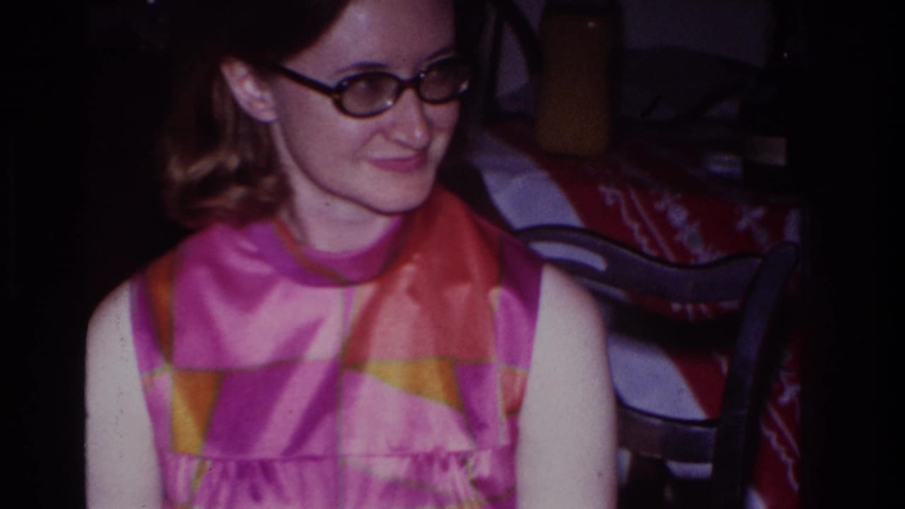 Young woman in glasses smiles slightly beside a patterned tablecloth and chairs.
