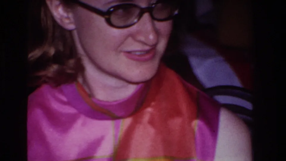 The 8mm frame moves into a tight close-up of the woman in glasses, showing her dress pattern and soft indoor lighting.