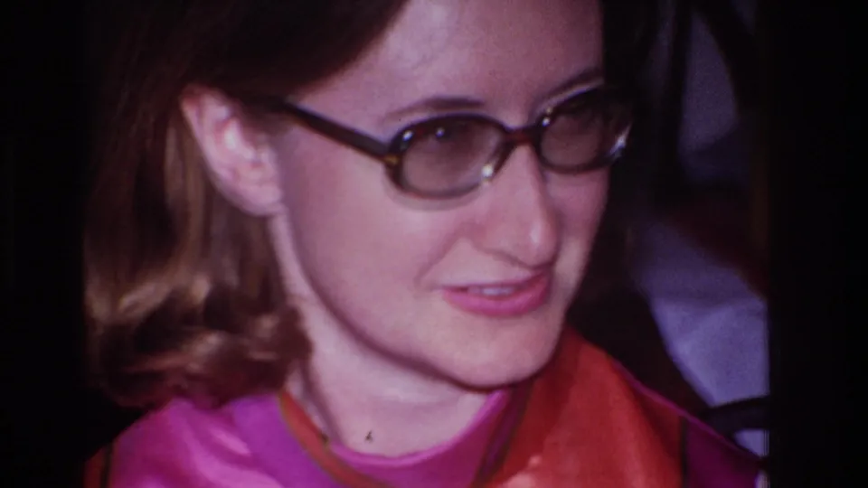 A close 1967 home-movie frame shows the woman in glasses looking down and aside, with her patterned dress and interior background visible.