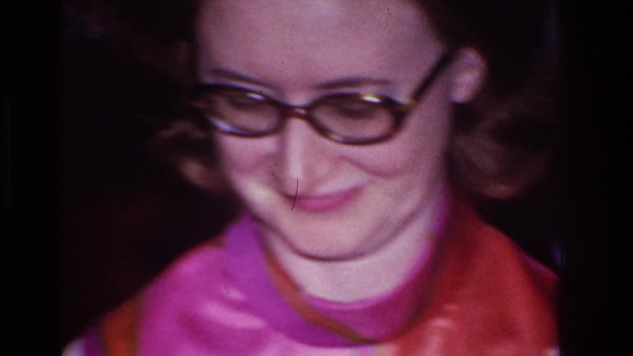 Woman in round glasses smiles while looking downward in dim indoor home-movie footage.