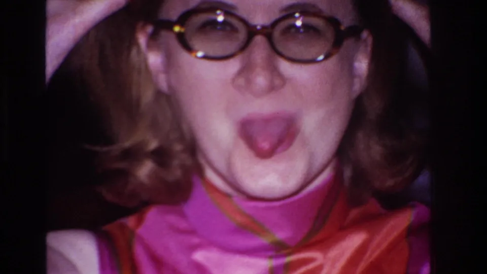 The woman in round glasses sticks out her tongue at the camera, with hands lifted near her head in a playful pose.