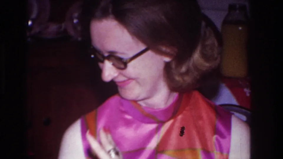 The woman turns in profile and smiles, with a wooden cabinet and bottles visible behind her in the indoor setting.