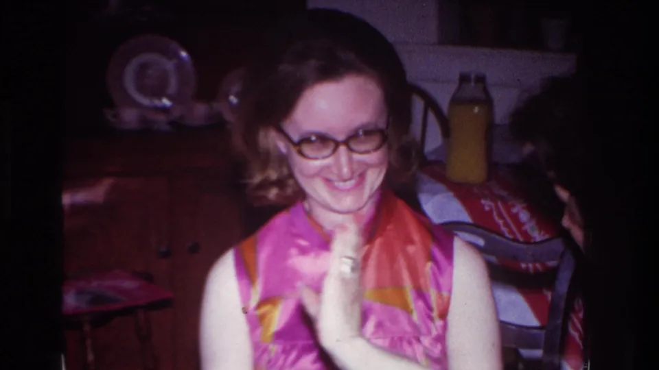 The woman smiles broadly with her hands together, seated or standing near a cabinet and household objects in the 1967 home-movie scene.
