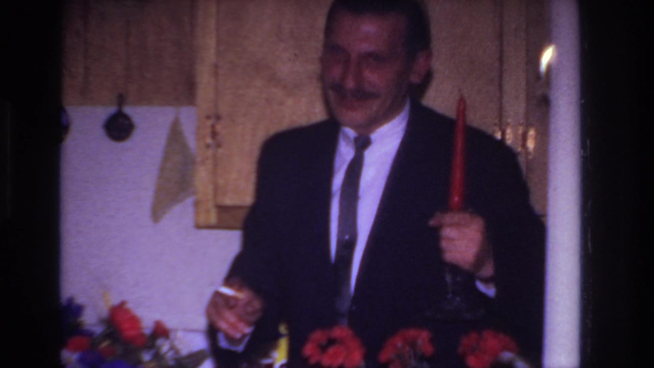 Mustached man in a dark suit holds a red candle and small glass in a kitchen.