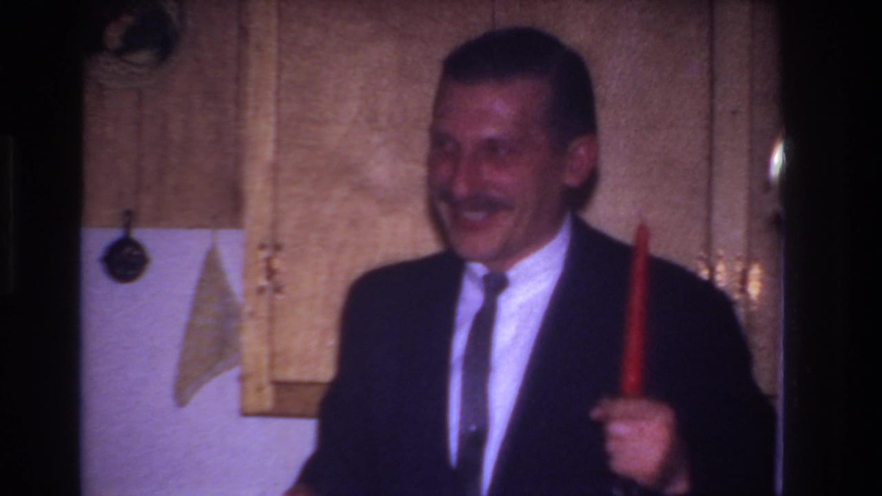 Smiling man in suit holds a tall red candle beside wood kitchen cabinets.