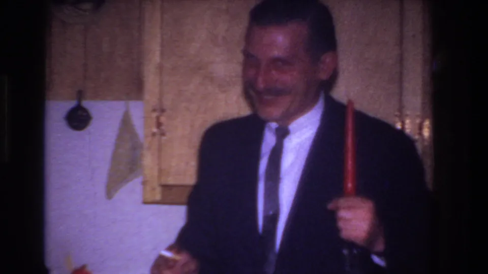 The suited man leans forward and smiles while holding the red candle, framed by cabinets and a white kitchen wall.