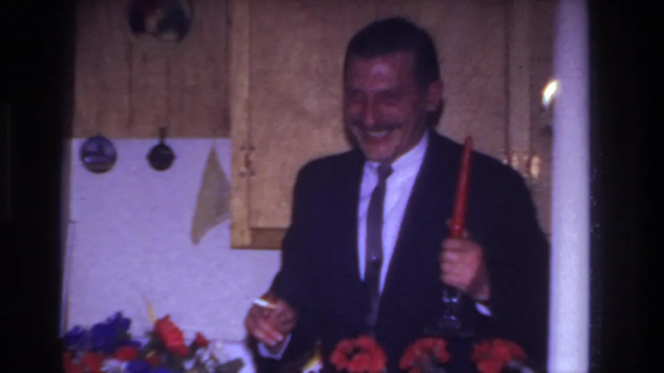 At the end of the 1967 home-movie clip, the man laughs while holding a red candle and glass in the kitchen.