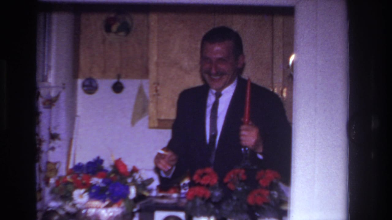 In a 1967 Rockaway Beach home movie, a suited man laughs while holding a red candlestick behind flowers in a kitchen.
