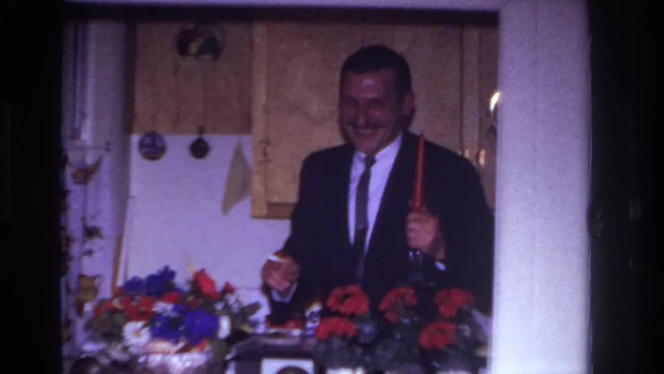 In a 1967 Rockaway Beach home movie, a suited man laughs while holding a red candlestick behind flowers in a kitchen.