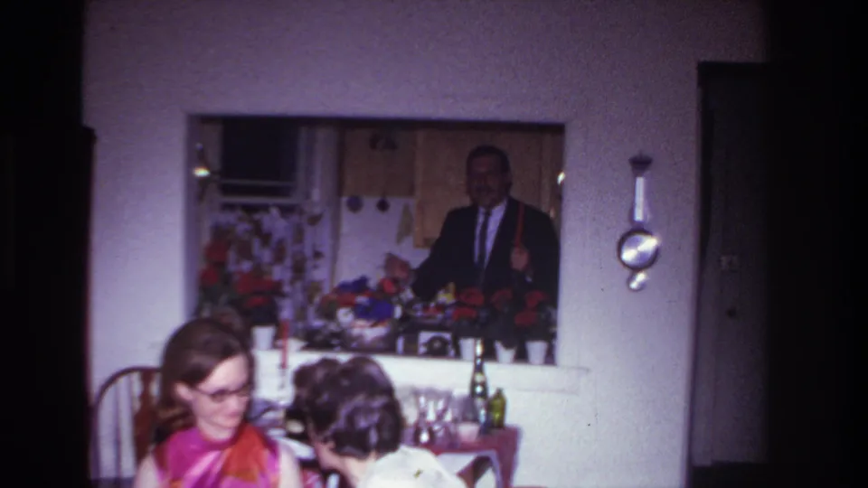 In the 1967 Rockaway Beach interior, two women sit at a table while a suited man stands in the kitchen background.