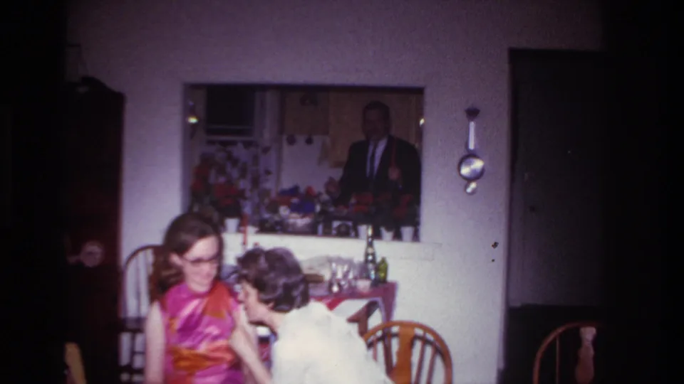 A 1967 Rockaway Beach home-movie frame shows two seated women at a party table with the suited man and flowers behind them.