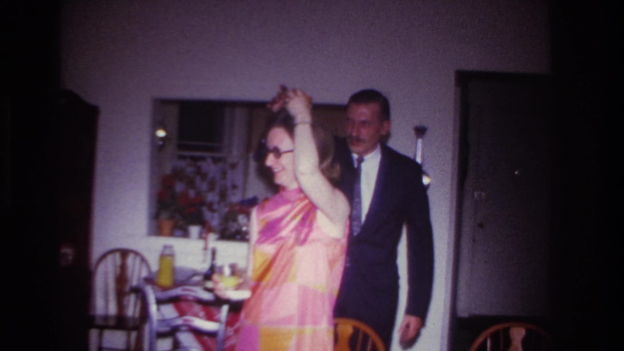 Woman in a bright patterned dress dances while a suited man stands behind her indoors.