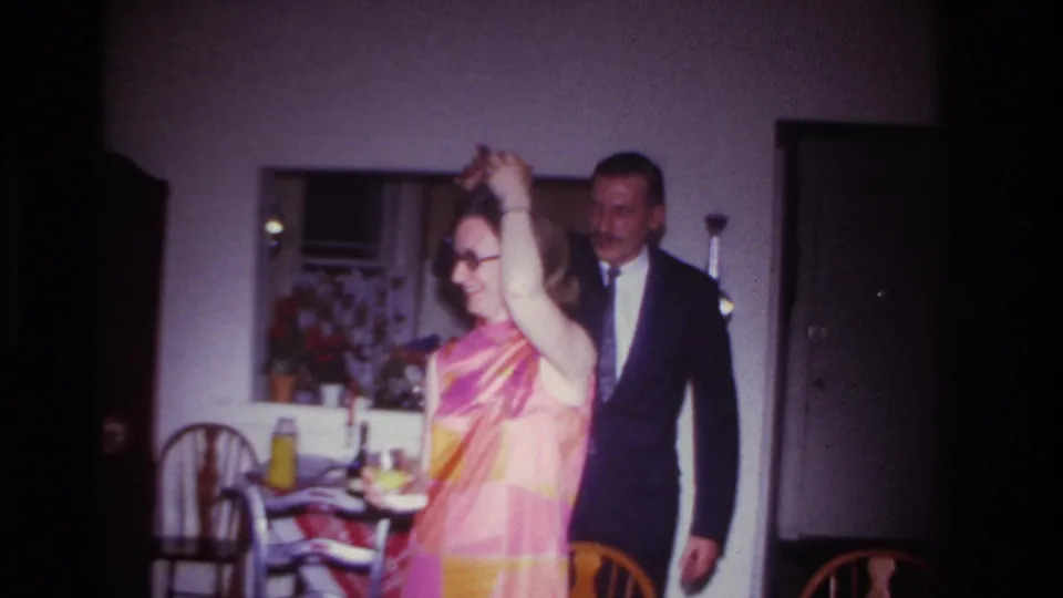 In a 1967 Rockaway Beach home-movie frame, a woman in a pink-orange dress raises her arms while a suited man stands nearby.