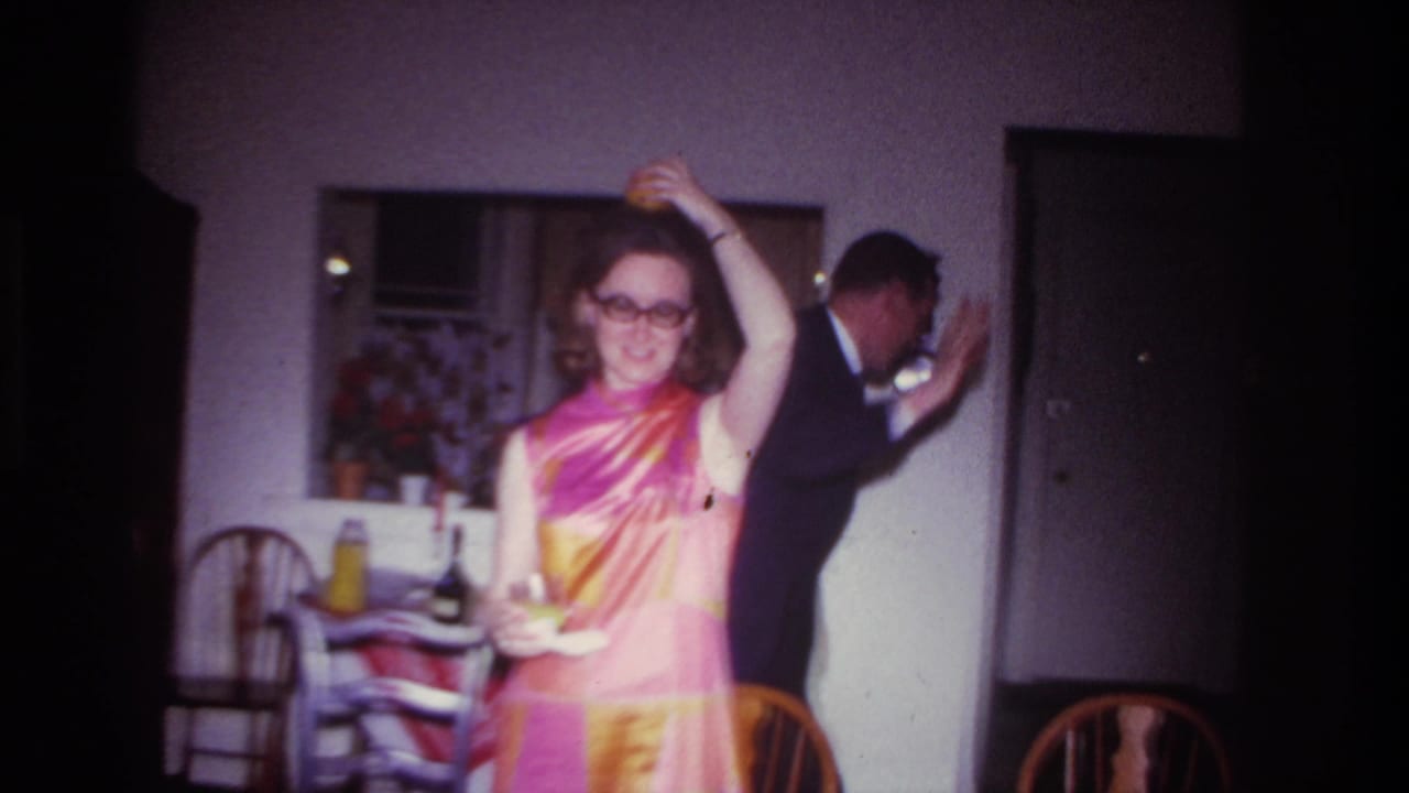 Woman smiles and dances with a drink while a suited man leans near a doorway.