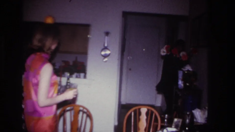 The woman turns across the room with a drink in hand, seen in a softly focused 1967 Rockaway Beach home-movie interior.
