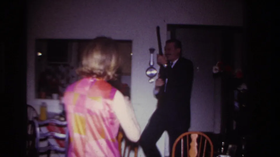 A woman in a patterned dress faces a suited man holding a long object, possibly a rod, inside the Rockaway Beach home setting.