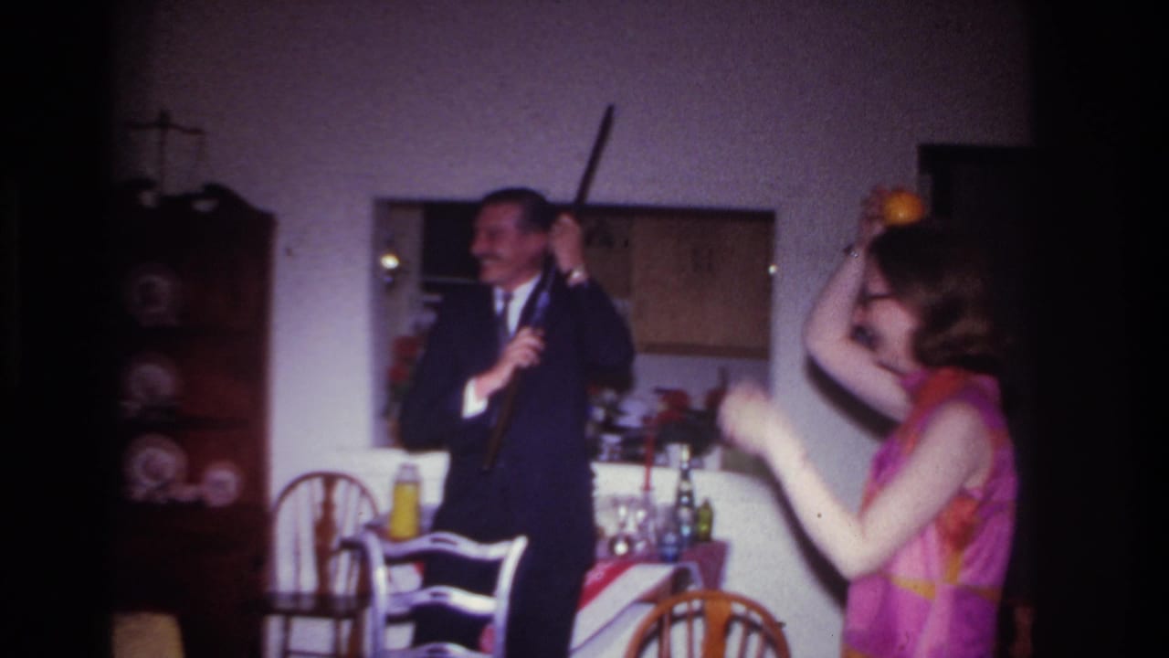 Suited man holds a long rifle-like object while a woman raises her arms nearby.