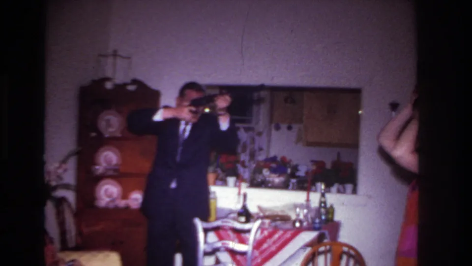 A 1967 home-movie frame shows a suited man posing with a long rifle-like object in a private dining room, with a china cabinet and party table visible behind him.