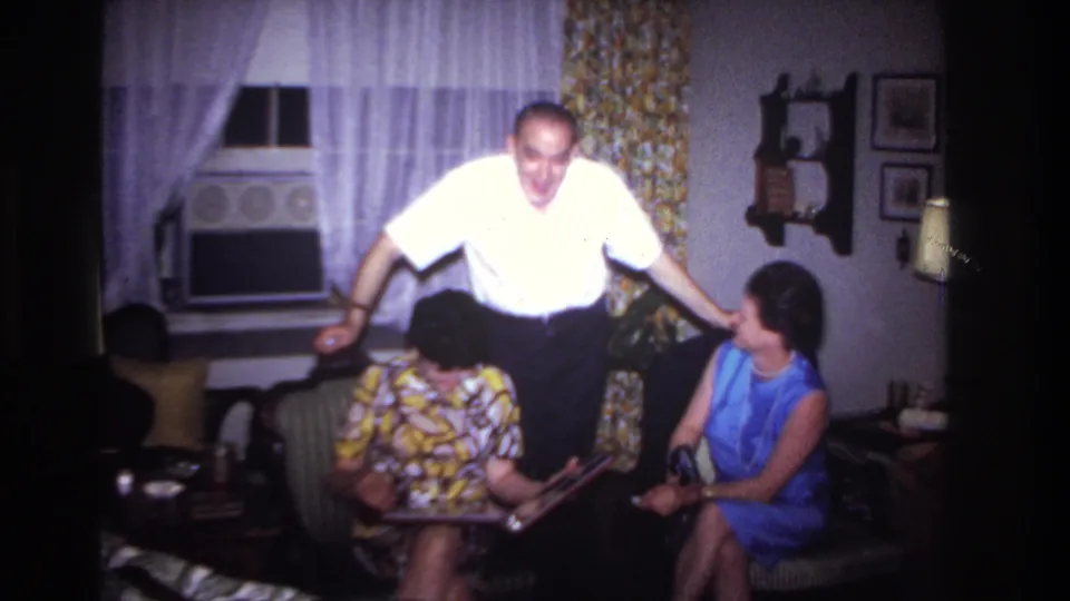 The clip shifts to a living-room moment in the Rockaway Beach home movie, where a man in a white shirt stands beside two seated women in patterned and blue dresses.