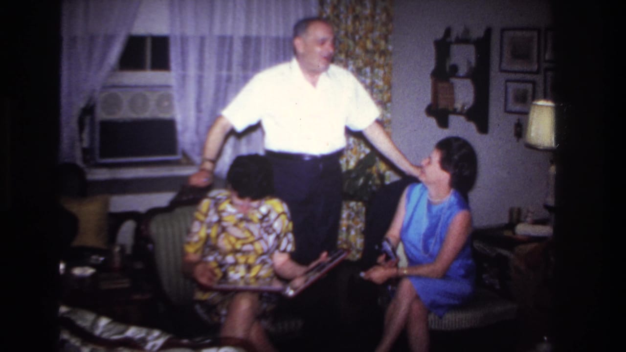 1967 Rockaway Beach home-movie frame showing a man in a white shirt standing beside two women seated with a photo album in a living room.