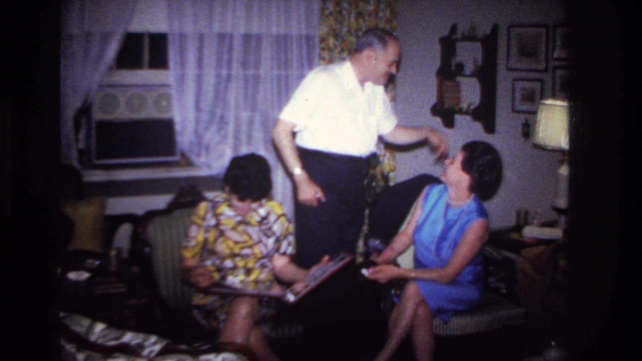 Man gestures toward a seated woman in blue while another woman looks down at a photo album.