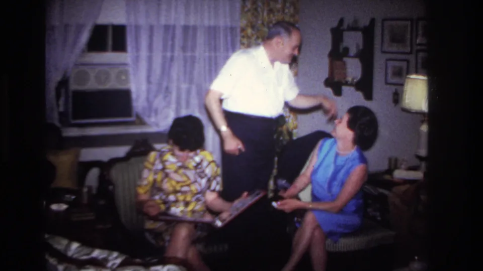 Rockaway Beach 1967 living-room frame with a standing man gesturing toward a seated woman in blue as another woman studies a photo album.
