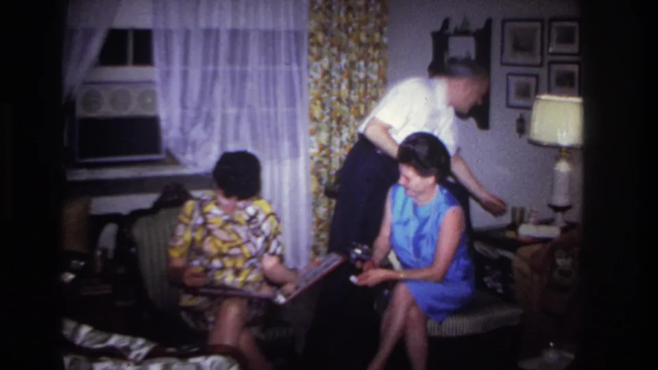 1967 home-movie frame from Rockaway Beach showing a man leaning close to two seated women as they handle a photo album indoors.