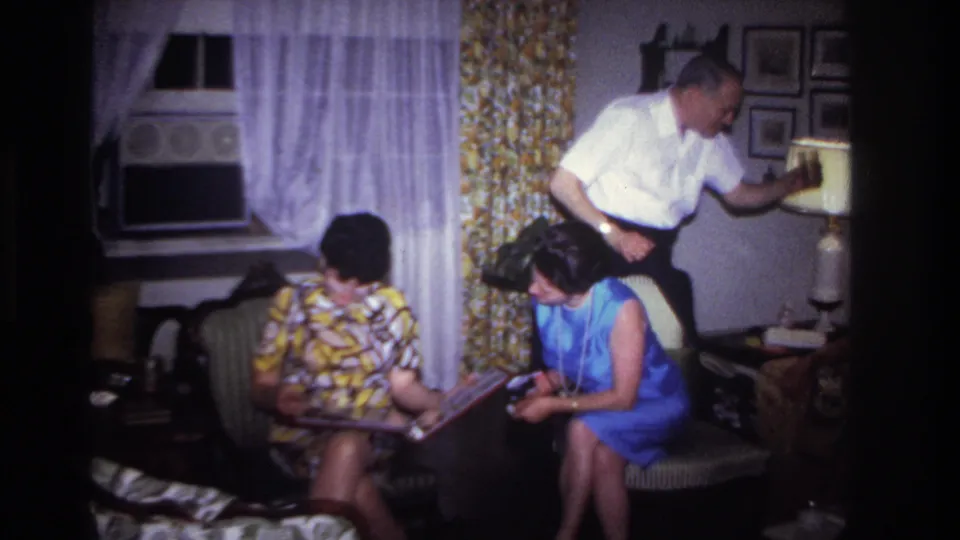 Rockaway Beach 1967 frame showing two women seated with a photo album as a man bends near a lamp in a private living room.