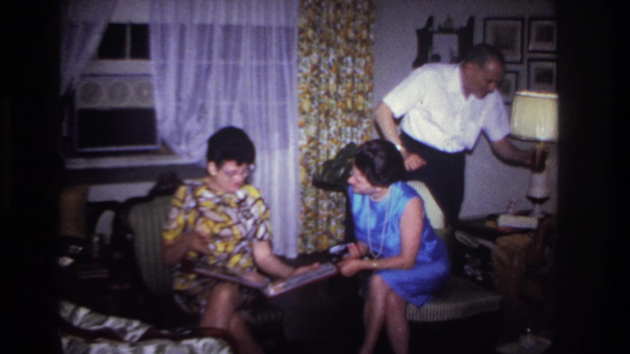 Two seated women look at a photo album while a man stands behind them in a living room.