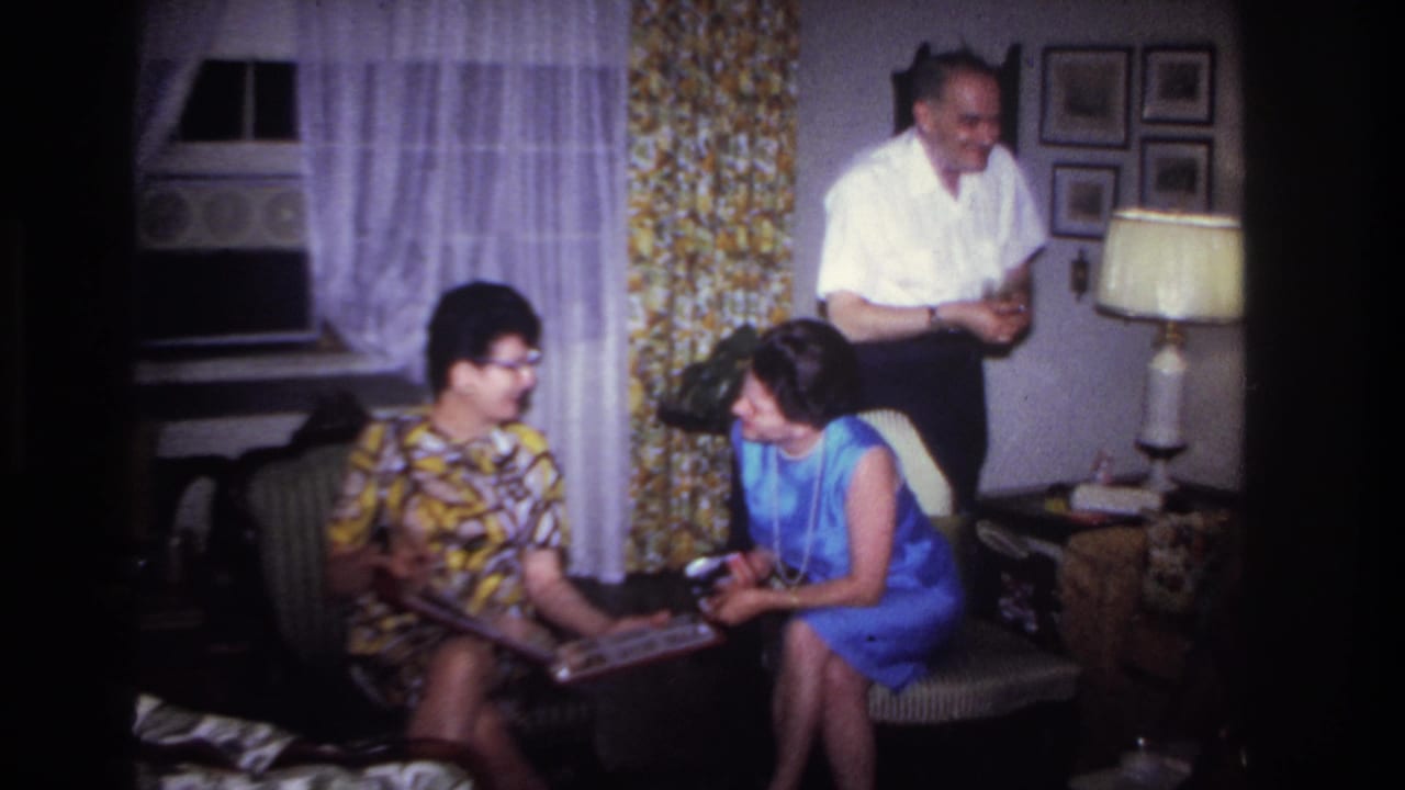 Two women examine a photo album in a Rockaway Beach home while a smiling man stands near the lamp and wall pictures.