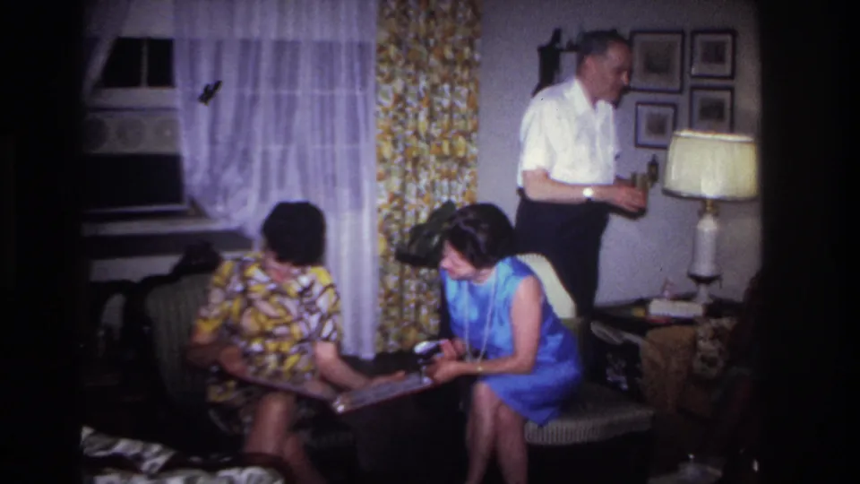 The home-movie frame shows two women focused on a photo album, with a man standing near floral curtains and a table lamp.