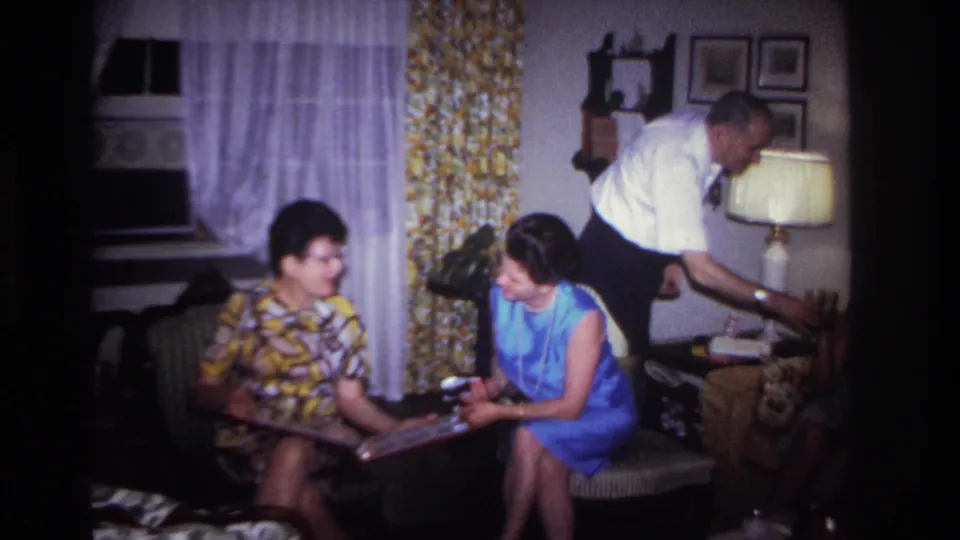 In the 1967 interior scene, two women sit with an album as the man turns toward a side table beside the lamp.