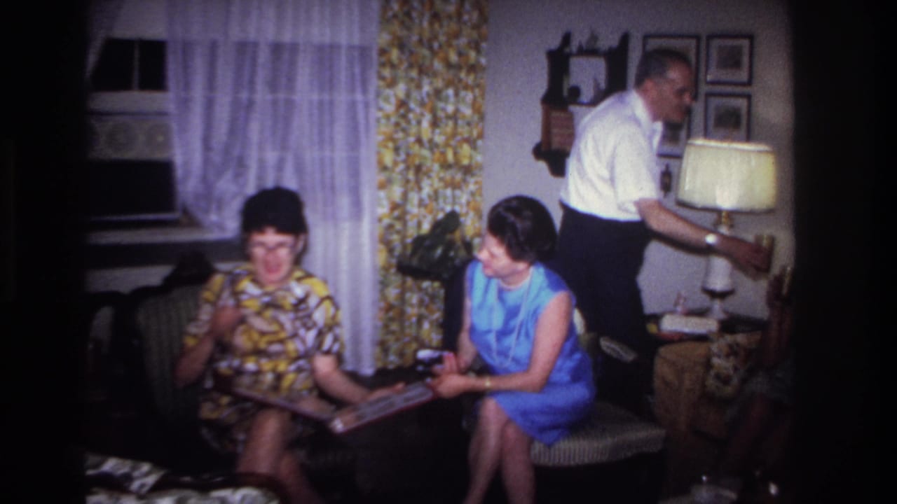 1967 Rockaway Beach home-movie frame of two women on a couch with a photo album as a man stands by a lamp.