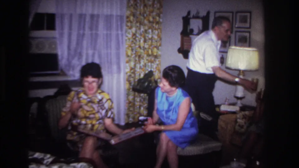 1967 Rockaway Beach home-movie frame of two women on a couch with a photo album as a man stands by a lamp.
