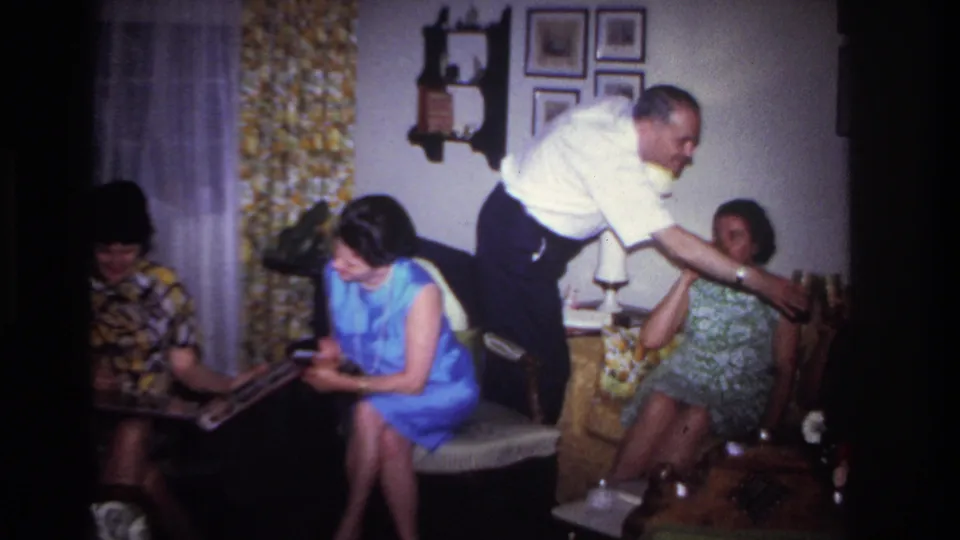 1967 Rockaway Beach home-movie frame of a man offering a drink near seated women in a decorated living room.