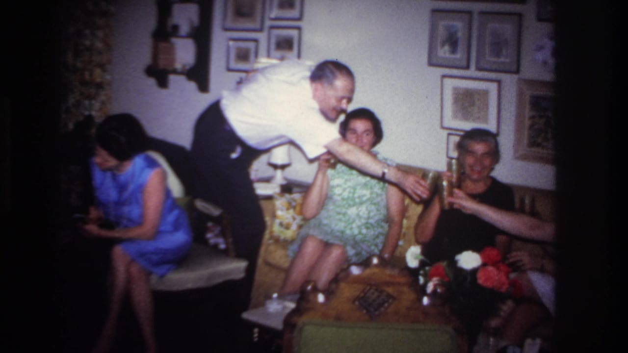 1967 Rockaway Beach home-movie frame showing adults on a yellow couch while a man in a white shirt serves drinks.