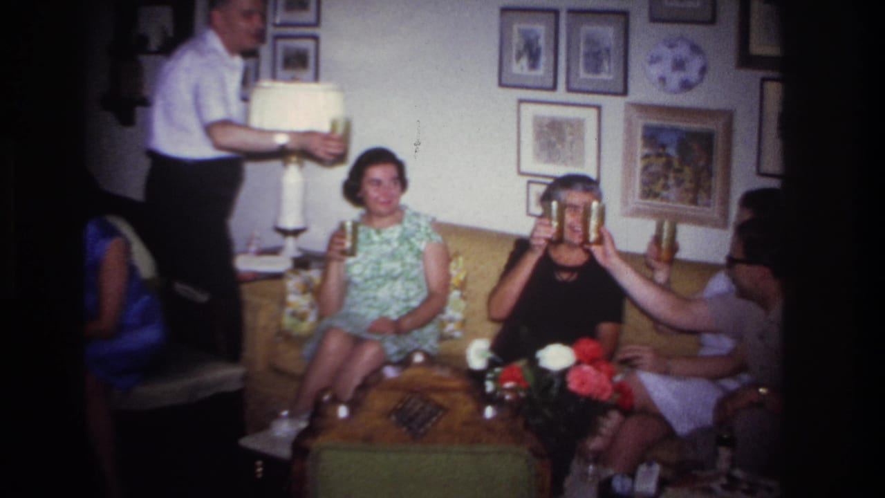 Seated women hold drinks while a man stands near a lamp in the living room.