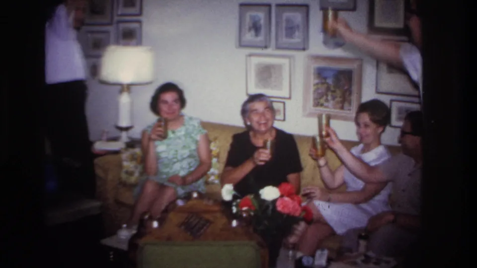 1967 Rockaway Beach home-movie frame showing women seated on a couch, smiling and raising glasses in a living room.