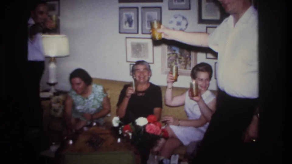 1967 Rockaway Beach frame of adults on a yellow couch raising drinks, with a man partly visible at the right edge.