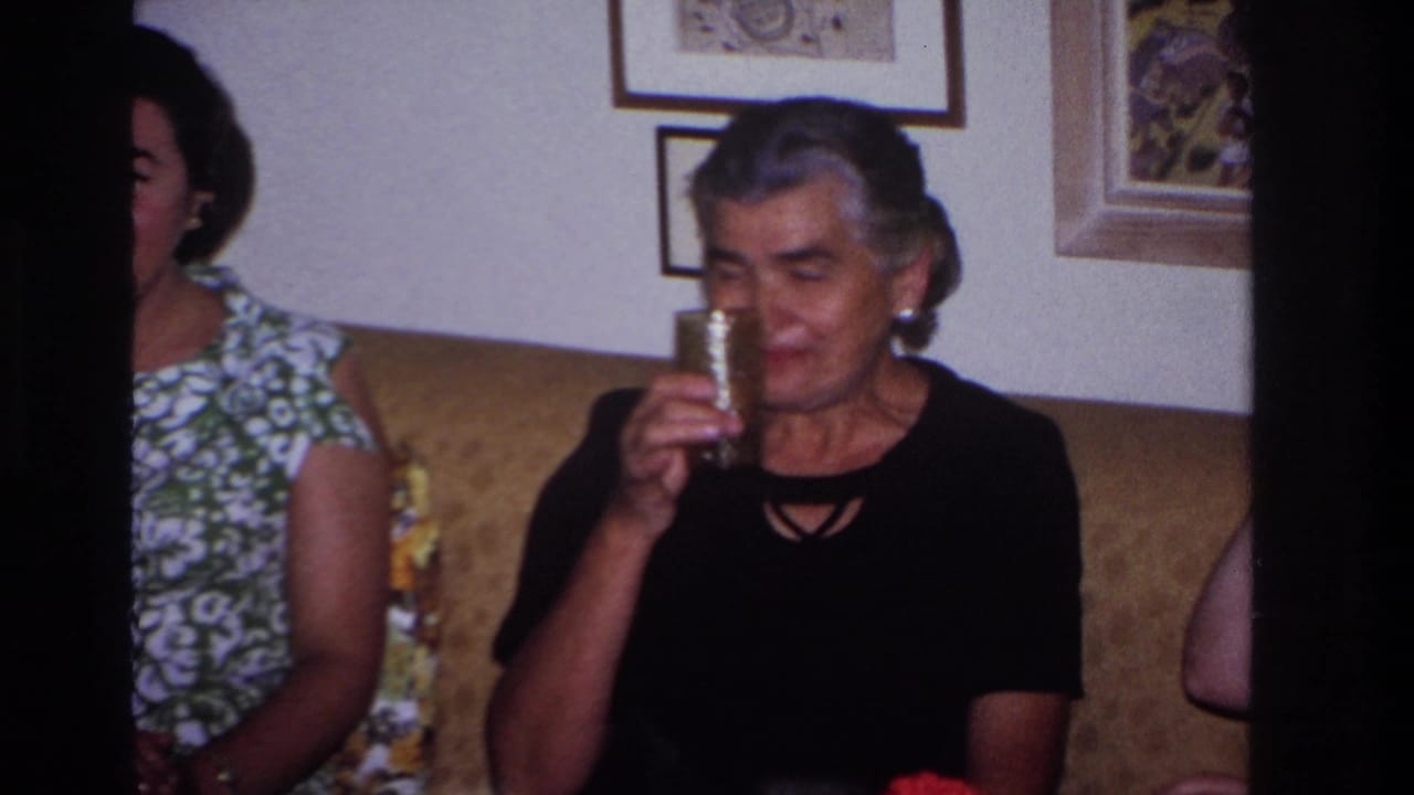 Women sit on a couch in a private living room, including an older woman in a black top holding a tall patterned glass.